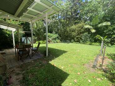 A shaded patio with a wooden dining table overlooks a bright, grassy backyard surrounded by lush green trees and plants, creating a peaceful, inviting outdoor space.