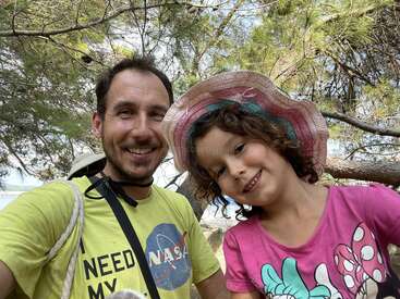 A smiling man and a girl pose outdoors among trees. The man wears a yellow NASA T-shirt; the girl wears a pink hat and Minnie Mouse shirt.