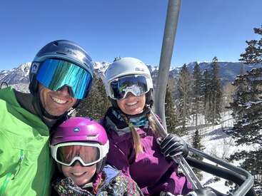 Une famille de trois personnes souriantes profite d'une journée ensoleillée à ski sur un télésiège, coiffées de casques et de lunettes de ski colorés, entourées de montagnes enneigées et de pins.