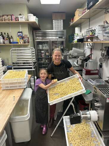 Une femme souriante et une jeune fille, toutes deux vêtues d'un tablier, tiennent fièrement des plateaux de pâtes fraîchement préparées dans une cuisine professionnelle remplie d'équipements, d'étagères et de provisions.