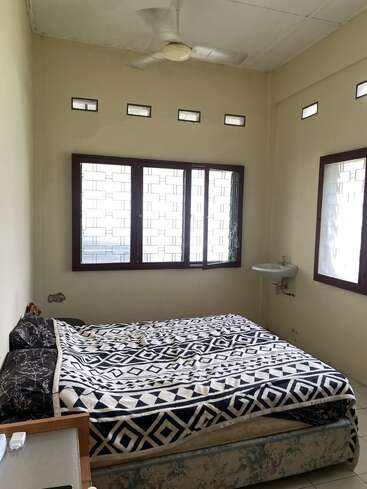 Une petite chambre simple avec une grande fenêtre, un lit avec des draps à motifs, un ventilateur au plafond, un petit lavabo et un sol carrelé blanc. Les murs sont percés de petites fenêtres.