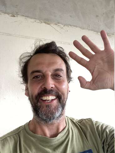 Un homme barbu et moustachu sourit et salue l'appareil photo. Il porte une chemise verte et se tient debout sur un fond uni.