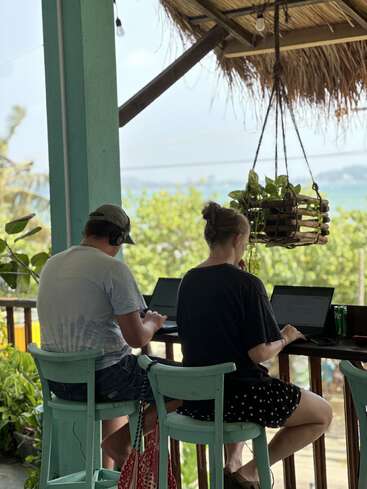 Duas pessoas estão sentadas em bancos turquesa em um balcão de madeira, trabalhando em seus laptops. Elas contemplam uma vista tropical e arborizada sob um telhado de palha, aproveitando um ambiente tranquilo.