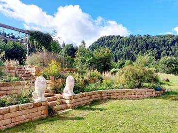 Ein wunderschöner Garten mit terrassenförmigen Steinmauern, zwei weißen Löwenstatuen, üppigen grünen Pflanzen, strahlend blauem Himmel und waldbedeckten Hügeln im Hintergrund. Friedliche Umgebung.