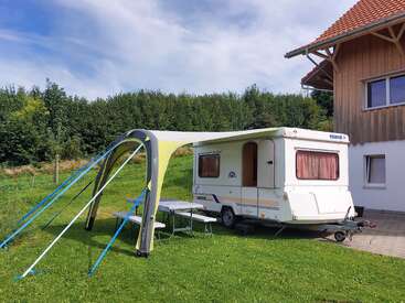 Ein kleiner weißer Wohnwagen ist neben einem Haus auf grünem Gras geparkt. Ein Sonnensegel mit blauen Seilen beschattet einen Picknicktisch und eine Bank. Bäume umgeben die Szene.