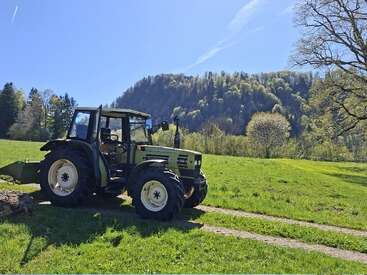 A green tractor is parked in a lush grassy field under a clear blue sky, with forested hills and scattered trees in the scenic background.