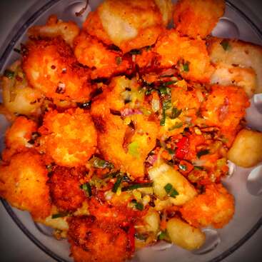 A bowl filled with crispy, golden-brown fried cauliflower florets, garnished with chopped green herbs, red chilies, and onions, giving it a vibrant, appetizing look.