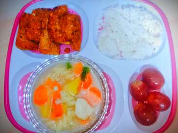 This image shows a compartment tray with rice, fried tempeh, grapes, and a bowl of vegetable soup. Bright colors indicate a balanced, nutritious meal.