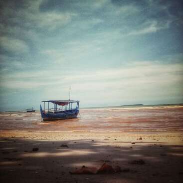 A blue boat rests on shallow, sandy waters near the shore under a partly cloudy sky. Distant boats and an island appear on the horizon. Peaceful scene.