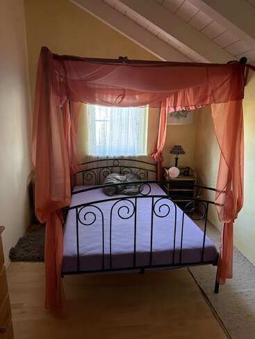 Una acogedora habitación con una cama con dosel de metal negro cubierta con una tela rosa transparente, ropa de cama lavanda, una pequeña ventana, una mesita de noche y un ambiente cálido y acogedor.