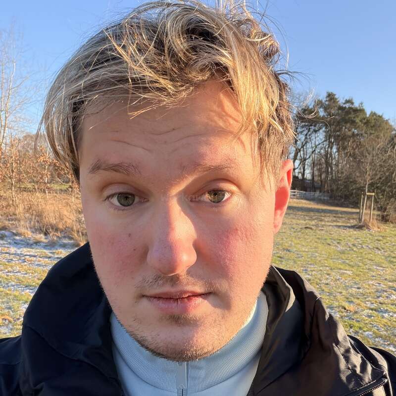 Un joven con el pelo rubio revuelto está de pie al aire libre en un día soleado y frío. Lleva una chaqueta negra, con hierba y árboles al fondo.
