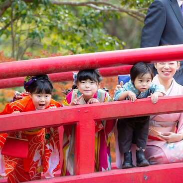 Three children in colorful traditional Japanese kimonos and one woman smile and pose on a red bridge, surrounded by greenery and autumn foliage, enjoying a festive outing.