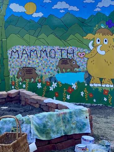Um mural colorido mostra um mamute de desenho animado, dois filhotes de mamute, montanhas, flores e a palavra “MAMMOTH”. À frente, há tijolos, toalhas e materiais de pintura.