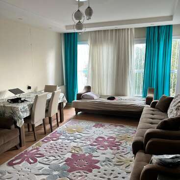 The image depicts a living room with a dining table, couch, and bed, featuring a floral rug, white and teal curtains, and a window with a view of trees outside.