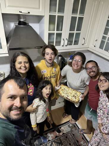 Sechs lächelnde Erwachsene und zwei aufgeregte Kinder versammeln sich in einer gemütlichen Küche und präsentieren fröhlich ein frisch gebackenes Gericht auf einem Tablett, bereit, eine Mahlzeit zu teilen.