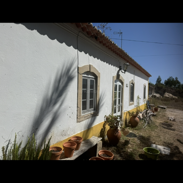 Uma casa rural branca com detalhes em amarelo e telhado de terracota, vasos de plantas e bancos de jardim. A luz do sol intensa projeta sombras; grama seca e árvores cercam o prédio.