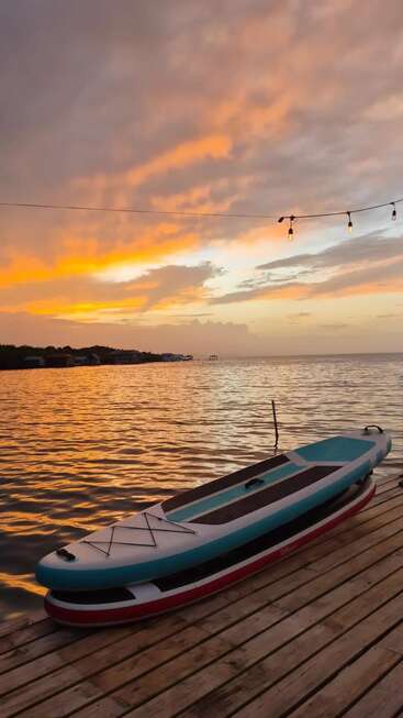 Un coucher de soleil apaisant teinte le ciel d'orange au-dessus d'une mer calme. Deux planches de paddle reposent sur un ponton en bois, surmonté de guirlandes lumineuses qui créent une atmosphère paisible.