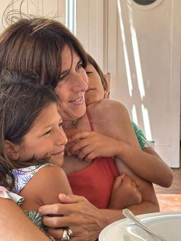 Eine Frau umarmt liebevoll zwei Kinder; alle lächeln herzlich und genießen diesen innigen Moment. Sonnenlicht strömt herein und schafft eine friedliche, fröhliche Familienszene im Haus.