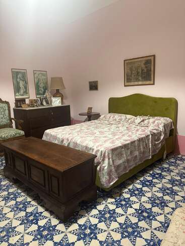 Dieses gemütliche Schlafzimmer besticht durch ein grün gepolstertes Bett, eine geblümte Tagesdecke, Holzmöbel, Vintage-Dekor, gerahmte Kunstwerke, einen gemusterten blau-weißen Fliesenboden und zartrosa Wände.