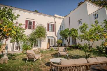Dieses Bild zeigt eine bezaubernde weiße Villa im mediterranen Stil mit roten Fensterläden, einem üppigen Garten, einer Sitzecke im Freien, Zitronenbäumen und einer friedlichen, sonnigen Atmosphäre unter klarem Himmel.
