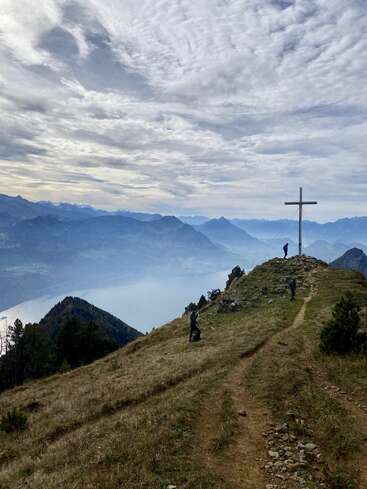 A winding mountain path leads to a summit with a large cross. Hikers stand nearby, overlooking misty valleys, distant mountains, and a dramatic cloud-filled sky.