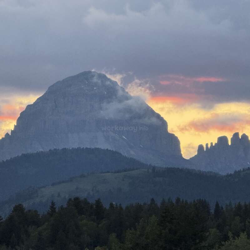 Una imponente montaña se alza majestuosa bajo unas nubes espectaculares. La puesta de sol baña el cielo de tonos anaranjados y amarillos, mientras densos bosques cubren las onduladas colinas.
