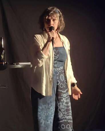 A woman stands on stage holding a microphone, illuminated by stage lights. She wears a patterned blue dress, white shirt, and appears to be performing stand-up comedy.