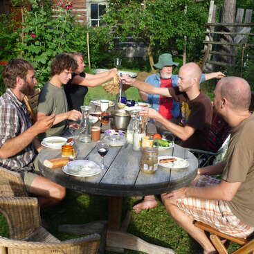 Das Bild zeigt eine Gruppe von sechs Personen, die um einen runden Holztisch in einem Garten versammelt sind und gemeinsam eine Mahlzeit genießen. Sie sitzen in Korbsesseln.