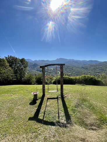 Eine Holzschaukel steht allein auf einem grasbewachsenen Hügel, das Sonnenlicht wirft lange Schatten und im Hintergrund sind die Berge, der blaue Himmel und die Bäume zu sehen.