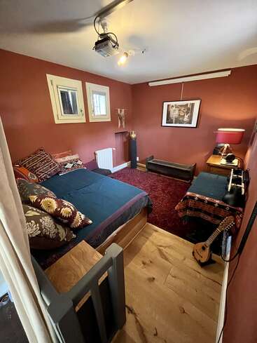 Gemütliches Schlafzimmer mit erdroten Wänden, Holzboden, Doppelbett, Zierkissen, Gitarre, gerahmter Kunst, Schreibtisch mit Lampe, Projektor und zwei kleinen Fenstern.