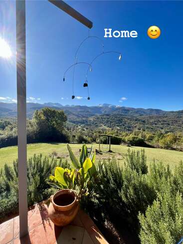 Eine friedliche Terrasse mit Blick auf einen üppigen grünen Garten und die Berge in der Ferne. Strahlender Sonnenschein, klarer blauer Himmel und das Wort "Zuhause" vermitteln Wärme und Ruhe.
