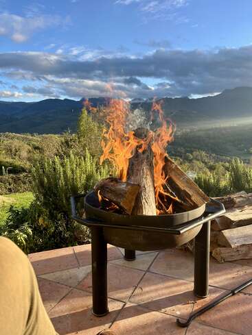 Eine Feuerstelle mit brennenden Holzscheiten steht auf einer Terrasse. Dahinter erstrecken sich üppiges Grün und Berge unter einem blauen Himmel mit vereinzelten Wolken. Eine friedliche Umgebung.
