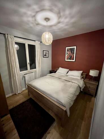 Dieses gemütliche Schlafzimmer verfügt über ein Doppelbett aus Holz, weiße Bettwäsche, zwei Nachttische mit Lampen, einen Teppich, Kunstwerke an einer roten Akzentwand und ein Fenster.