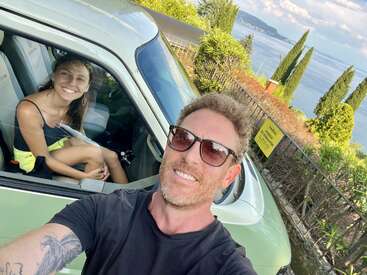Ein Mann macht ein Selfie im Freien neben einem grünen Auto. Darin sitzt eine lächelnde Frau. Hinter ihnen sind üppiges Grün, ein Zaun und ein malerischer Blick auf das Wasser zu sehen.