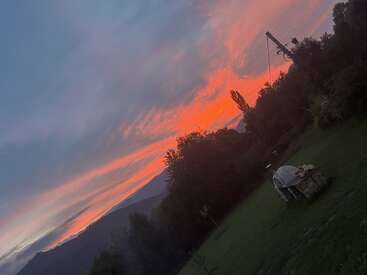 Ein dramatischer Sonnenuntergang füllt den Himmel mit orangefarbenen und rosafarbenen Schlieren. Bäume und Hügel umrahmen ein grünes Feld mit einem überdachten Steinofen.