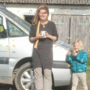 A woman and a child stand smiling in front of a white van. Both hold cups. A pumpkin rests on the ground. Sunlight brightens the scene.