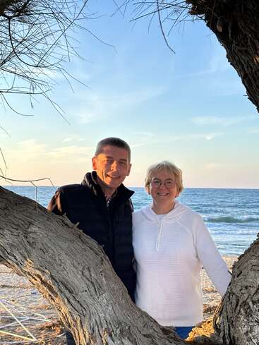 Un couple souriant se tient debout, encadré par des branches d'arbres, sur une plage au coucher du soleil, avec les vagues calmes de l'océan et un ciel bleu en toile de fond sereine.