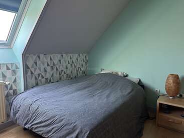 Cette chambre confortable se caractérise par un plafond en pente, un mur d'accent aux motifs géométriques, une lucarne, un lit double avec une parure de lit grise, une table de chevet, une lampe en osier et des murs vert menthe.