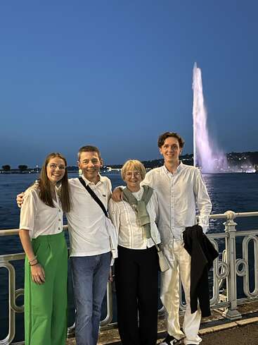 Quatre personnes sourient, debout près d'une balustrade au bord de l'eau, à la tombée de la nuit. Derrière elles, une grande fontaine illuminée projette un jet d'eau vers le ciel. Elles ont l'air heureuses, vêtues de tenues légères.