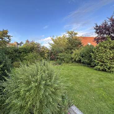 Un jardin verdoyant, parsemé d'arbustes et d'arbres, entoure une pelouse soigneusement tondue. Le ciel est partiellement nuageux, et un bâtiment au toit rouge se profile à l'horizon.