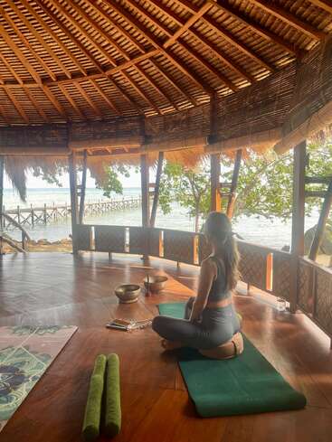 L'image représente une scène sereine d'une femme méditant sur un tapis de yoga dans une cabane au bord de la mer, entourée d'éléments apaisants et d'une vue paisible sur l'océan.
