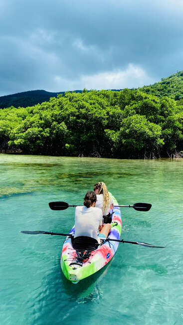 L'image représente deux personnes pagayant un kayak vibrant sur un plan d'eau serein, entouré d'une verdure luxuriante et de montagnes majestueuses au loin.