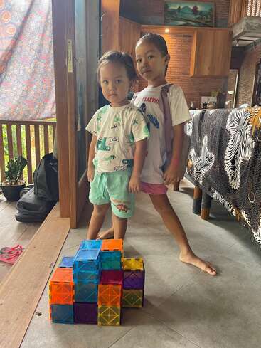 Deux jeunes enfants se tiennent avec assurance à l'intérieur, à côté d'une structure colorée construite à partir de carreaux magnétiques translucides. La pièce présente des murs en bois, des tissus à motifs et une lumière naturelle intense.