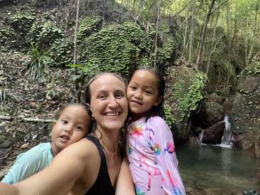 Une femme et deux enfants posent joyeusement pour un selfie devant une petite cascade, entourée de verdure luxuriante et de rochers dans un cadre forestier paisible.