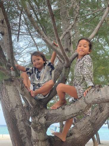Deux enfants souriants sont assis pieds nus sur des branches d'arbre au bord de la plage, entourés d'aiguilles de pin vertes et d'une eau bleue limpide, profitant d'un moment de jeu et de joie ensemble.
