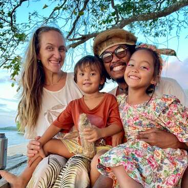 A happy family of four sits outdoors, smiling warmly. They are surrounded by nature, under a tree, with a blue sky and calm water behind them.