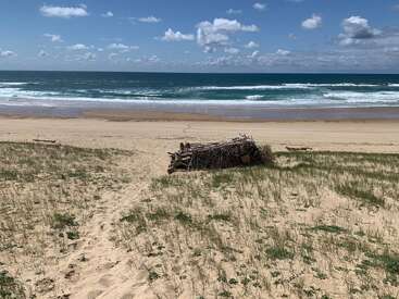 Uma praia de areia se estende até o oceano, com um pouco de grama aqui e ali. No meio, tem um abrigo feito de madeira flutuante, com ondas e um céu azul meio nublado ao fundo.