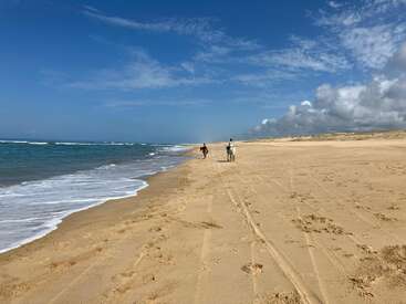 Uma enorme praia de areia se estende sob um céu azul. Duas pessoas, uma delas montada em um cavalo, caminham pela orla. Ondas suaves batem na areia intocada.