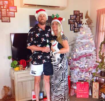 Um casal com chapéus festivos está ao lado de uma árvore de Natal branca, segurando um cachorro. Presentes, enfeites e fotos de família criam um clima alegre de fim de ano em casa.