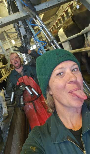 Duas pessoas numa sala de ordenha fazem caretas, deitando a língua para fora. Elas estão vestidas com roupas de trabalho e cercadas por vacas e equipamentos de ordenha.